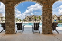 Inviting view of a luxurious apartment community pool framed by elegant stone arches, featuring comfortable lounge chairs under a bright blue sky.